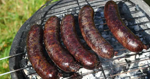 Pięć ciemnych, rumianych kiełbasek na ruszcie grilla. Widać dymek i żar, co sugeruje, jak powstaje kaszanka.