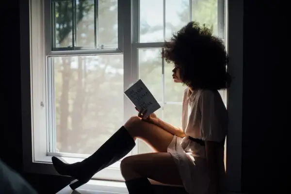 Vrouw met afro, zittend bij het raam met een boek, diep nadenkend over haar relatie.
