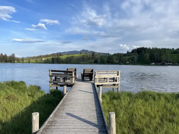 Holzsteg mit Sitzgelegenheiten am bayerischen See, umgeben von grünen Wiesen und bewaldeten Hügeln unter blauem Himmel.