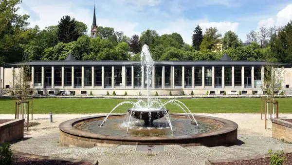Ein Brunnen sprudelt vor einem modernen Gebäude mit Säulen und Glasfronten. Im Hintergrund ein Kirchturm und Bäume. Ein idyllischer tschechischer Kurort.