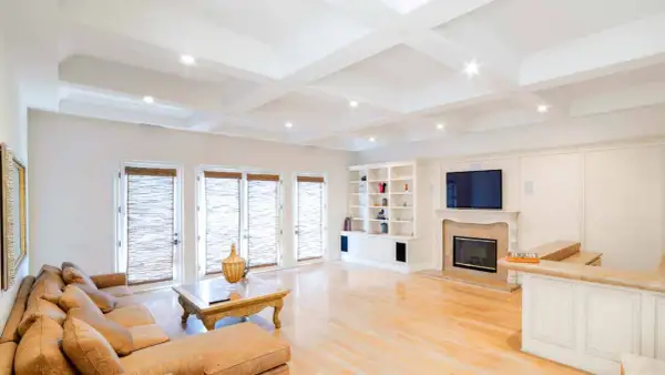 A spacious living room with a coffered ceiling featuring eight can lights. A large sectional sofa faces a fireplace and entertainment center.