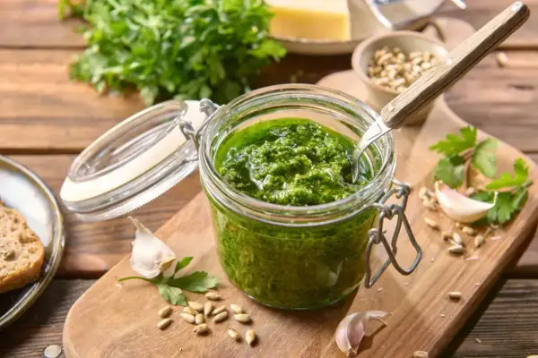Słoik domowego pesto z pietruszki, obok czosnek, nasiona słonecznika i kromka chleba.