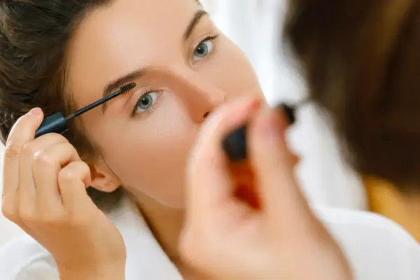 Une femme applique du mascara sur ses sourcils, préparant son maquillage bagage cabine pour un voyage.