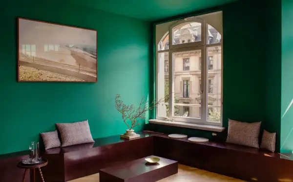 Un salotto con pareti verde scuro, un quadro paesaggistico, sedute in legno scuro e cuscini a quadri. I colori da abbinare al verde scuro includono il marrone del legno e le sfumature neutre dei cuscini.
