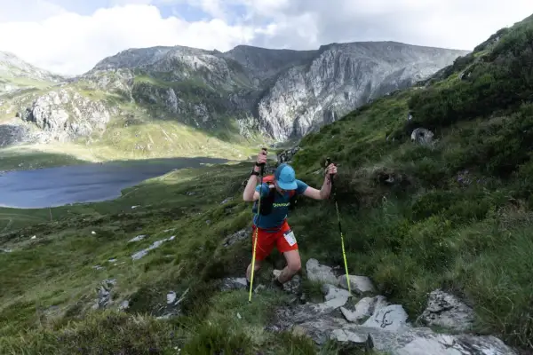 Un coureur grimpe une pente rocheuse, bâtons en main, lors de la kat100 by utmb. Un lac et des montagnes majestueuses en arrière-plan.