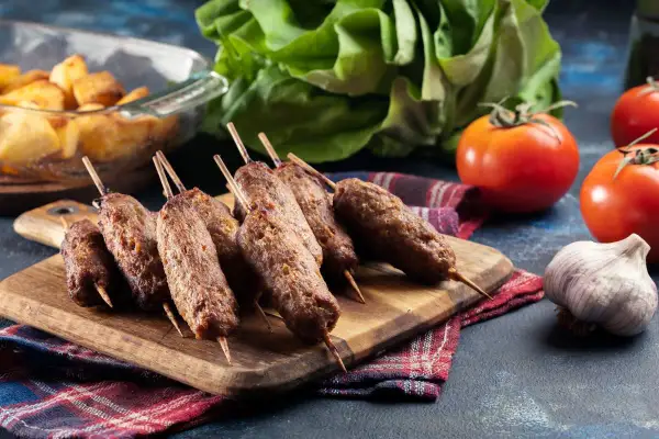 Aksamitne cevapcici z grilla, podane na desce z dodatkami: pieczone ziemniaki, pomidory i czosnek.