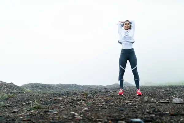 Femme en tenue de sport s'étirant sur un terrain rocailleux, prête pour son entrainement course à pied dans un paysage brumeux.