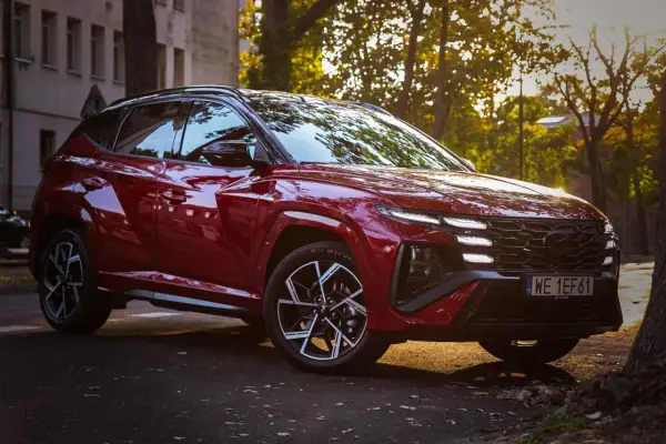 Czerwony Hyundai Tucson 2023 z czarnymi felgami i przyciemnianymi szybami, zaparkowany na tle kamienicy i drzew w jesiennym słońcu.