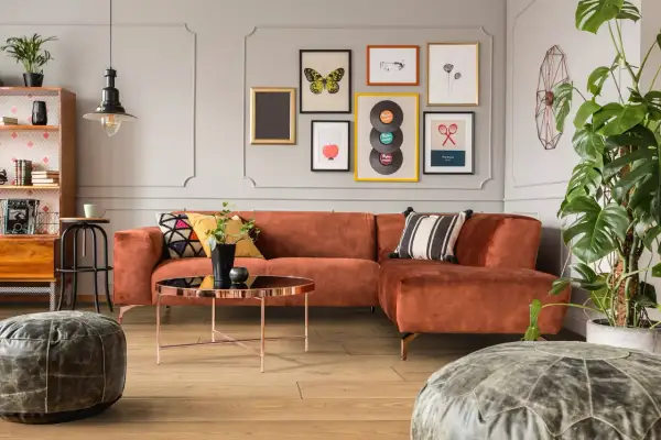 Pomarańczowa kanapa i stolik kawowy w stylu mid century modern. Na ścianie galeria obrazów z motywami retro.