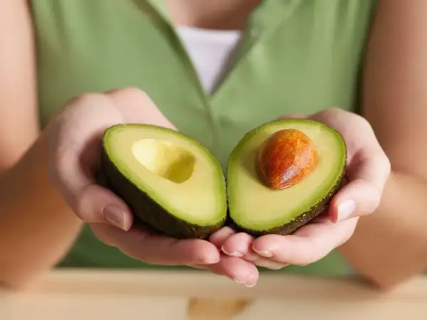 Manos sostienen un aguacate cortado por la mitad, mostrando su pulpa verde y el hueso. Así es qué es el aguacate, una fruta cremosa y nutritiva.