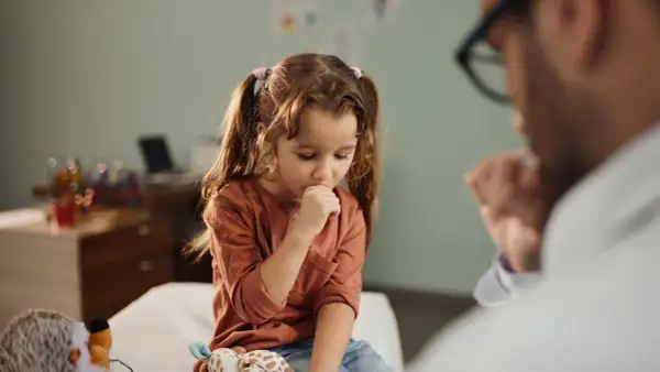 Niña con tos perruna en consulta médica. Un doctor la examina mientras ella se cubre la boca.