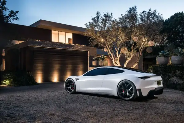 Biały roadster Tesla stoi na żwirowym podjeździe przed nowoczesnym domem.