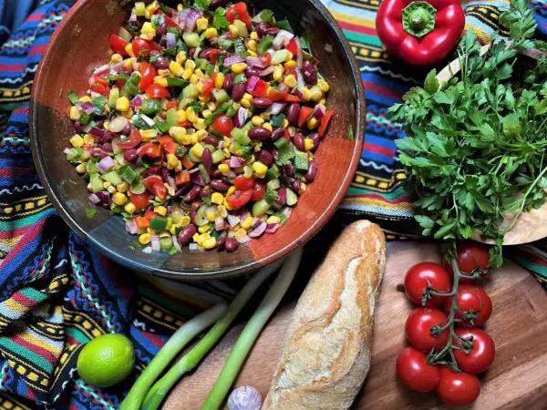 Bunter mexikanischer Salat mit Mais, Bohnen und Gemüse in einer Schüssel, daneben Baguette, Limette und Kräuter.