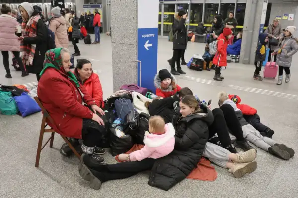Rodzina z dziećmi, w tym niemowlę, siedzi na podłodze z bagażami, czekając na pomoc. Ich status uchodźcy jest widoczny w ich sytuacji.