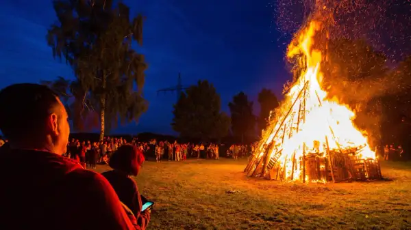 Menschen versammeln sich, um die Sommersonnenwende zu feiern. Ein riesiger Holzstapel brennt lichterloh im Abendhimmel, Funken steigen auf.