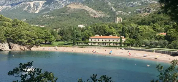 Malownicza zatoka z plażą w Czarnogórze. Woda jest turkusowa, a plaża piaszczysta, z kilkoma osobami odpoczywającymi pod parasolami. W tle widać zielone wzgórza i elegancki budynek.