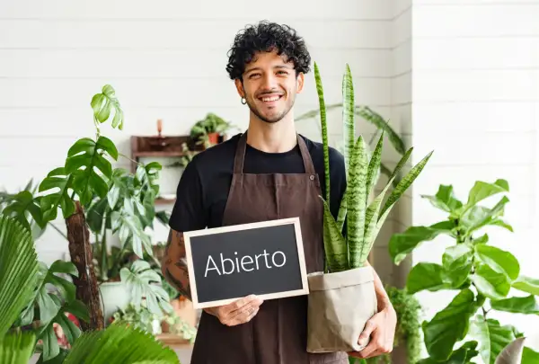 Joven sonriente con plantas, sosteniendo un cartel de "Abierto". Inspiración para ideas de negocios para jóvenes en el sector verde.