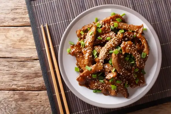 Pyszny kurczak teriyaki z sezamem i szczypiorkiem na talerzu, obok pałeczki.