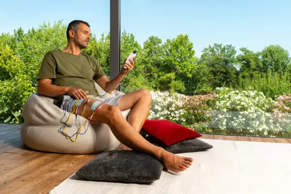 Homme assis, appliquant des électrodes sur sa cuisse, appareil de stimulation en main. Il observe le paysage fleuri, sans effets secondaires apparents du urostim.