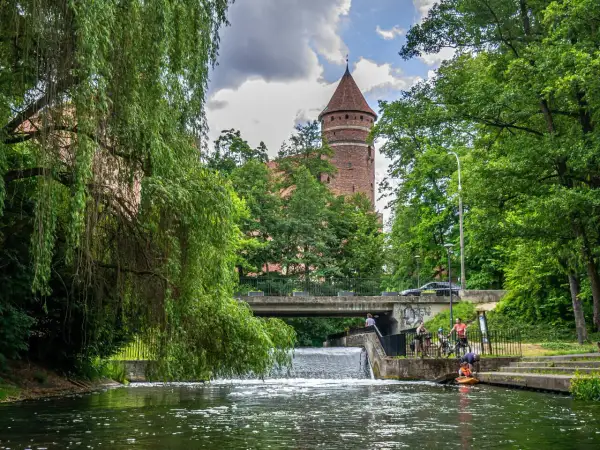 Malownicza przystań w Olsztynie z wieżą zamkową w tle. Woda, zieleń i ludzie odpoczywający nad rzeką tworzą spokojny krajobraz.