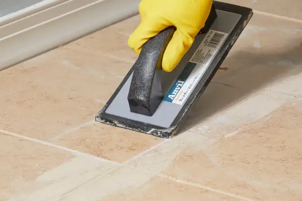 A gloved hand uses a grout float to spread grout, leaving a light grout haze on the tiled surface.