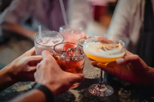 Ręce wznoszą kieliszki z kolorowymi drinkami. Wśród nich widać czerwony koktajl z lodem, różowy napój ze słomką i żółty drink w kieliszku. To idealny moment na toast z dobrym **medoc alkohol**!