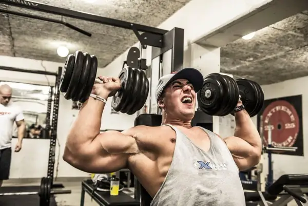 Man med keps och linne lyfter tunga hantlar i ett gym. Han pressar sig till max med varje repetition i sitt cluster set.