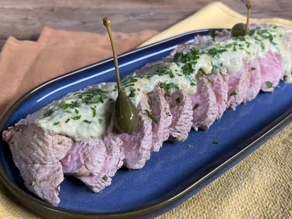 Tranches de vitello tonnato, nappées d'une sauce crémeuse et garnies de câpres et de persil, sur un plat bleu.