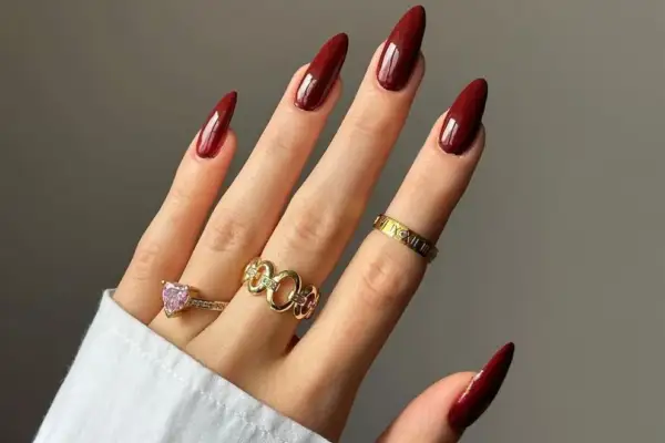 Main d'une femme aux ongles longs et pointus, vernis d'un bordeaux profond, parfaite tendance ongle automne. Bagues dorées et une bague cœur rose ornent ses doigts.
