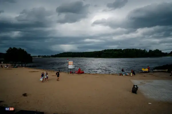 Plaża nad jeziorem Wysoka, z ciemnymi chmurami na niebie. Ludzie spacerują po piasku, niektórzy odpoczywają. W tle las i spokojna tafla wody.
