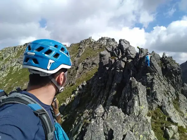 Alpinista w niebieskim kasku patrzy na grań Tatr, gdzie wspina się grupa ludzi.