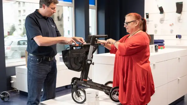 Mann erklärt einer Frau einen Rollator. Eine medizinische Hilfsmittel Liste ist im Hintergrund zu sehen.