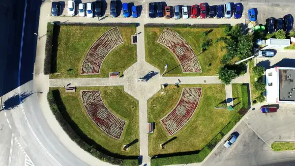 Piątek miasto środek Polski. Park miejski z klombami w kształcie łuków, alejkami i ławkami. W tle parking z samochodami.