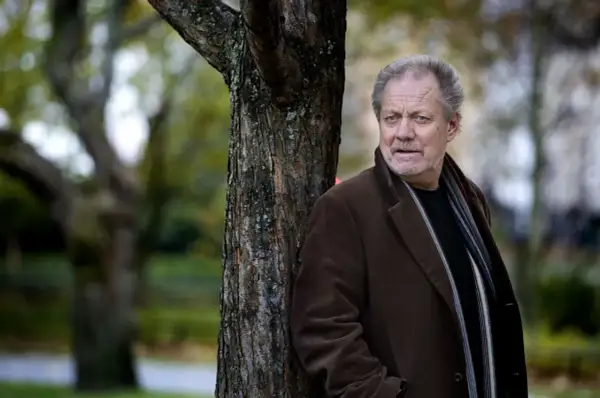 Harald Bovallius lutar sig mot ett träd i en park, klädd i brun rock och halsduk.