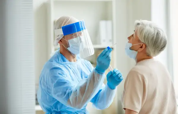 Arzt in Schutzkleidung nimmt bei älterer Frau einen Rachenabstrich. Testet man so auch auf MRSA, was ist das?