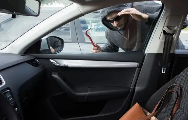 Un homme en capuche et lunettes de soleil tente un vol à la roulotte, outil rouge à la main, regardant à l'intérieur d'une voiture.