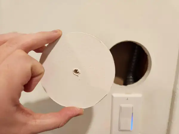 Un disco di cartongesso viene tenuto in mano vicino a un buco nel muro, con un interruttore della luce visibile sotto.