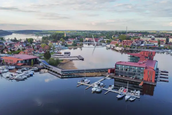 Widok na nowoczesny hotel i przystań jachtową na Mazurach. Widać liczne łodzie i żaglówki, a w tle miasteczko z charakterystyczną architekturą. To idealne mazury atrakcje dla miłośników żeglarstwa i wypoczynku nad wodą.