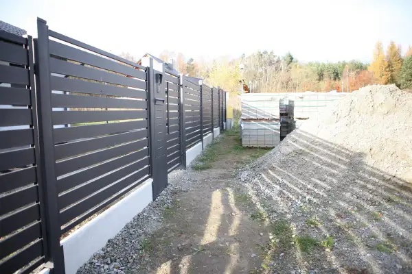 Schemat fundamentu pod bramę przesuwną. Widać nowoczesne ogrodzenie, materiały budowlane i hałdę żwiru.
