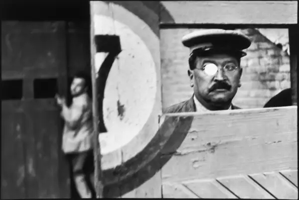 A man with a monocle and mustache, reminiscent of Cartier Bresson paintings, peers from behind a wooden structure. A blurred figure stands in the background.