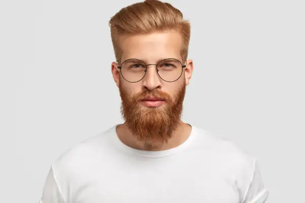 Un homme au visage rectangulaire, barbe rousse et lunettes rondes, porte un t-shirt blanc.