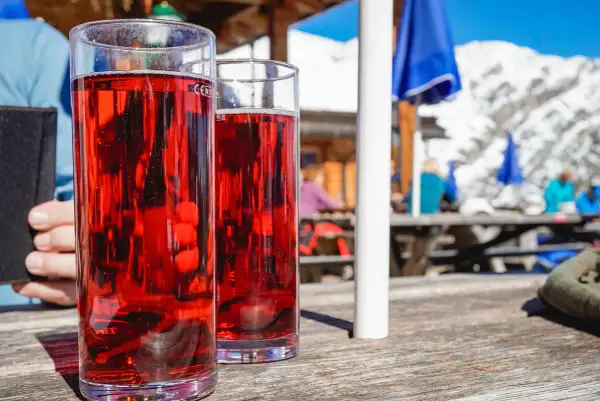 Zwei Gläser mit rotem Getränk, vielleicht ein Skiwasser-Rezept, stehen auf einem Holztisch. Im Hintergrund eine Skihütte mit Gästen.