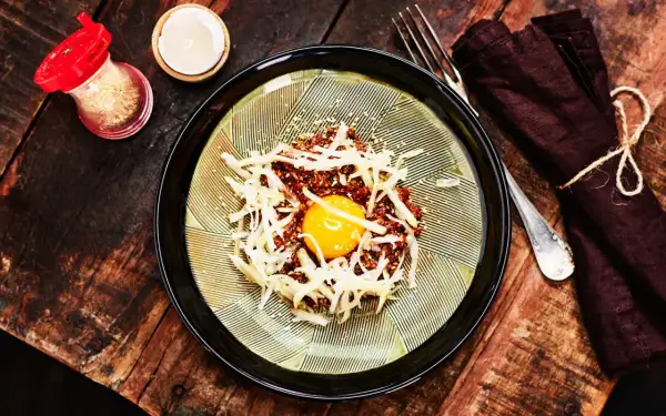 En läcker koreansk råbiff med en glänsande äggula i mitten, toppad med strimlad rättika och sesamfrön. Serveras på en vacker tallrik med en mörk servett och gaffel.