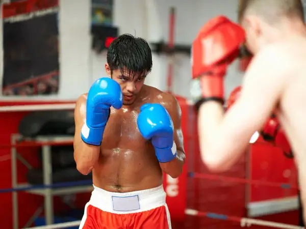 A boxer in blue gloves sweats during a tough boxing cardio workout in the ring, facing an opponent in red gloves.