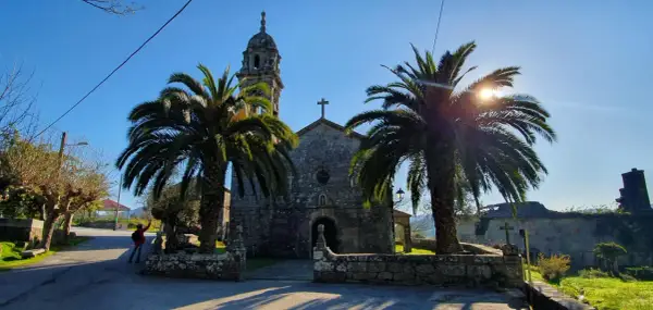 Iglesia de piedra con palmeras y sol brillante. Un lugar tranquilo que ver en Rianxo, ideal para pasear.