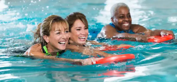 Three women joyfully participate in an adult learn to swim class, holding red kickboards as they move through the water.