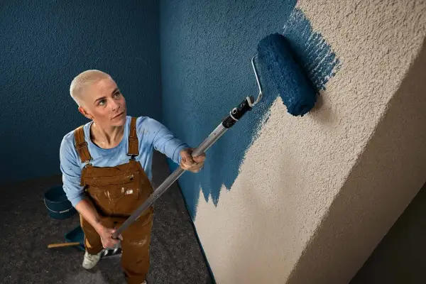 Frau mit Kurzhaarschnitt streicht mit einer Farbrolle eine Wand mit Rauputz. Sie trägt Latzhose und blaue Langarmshirt.