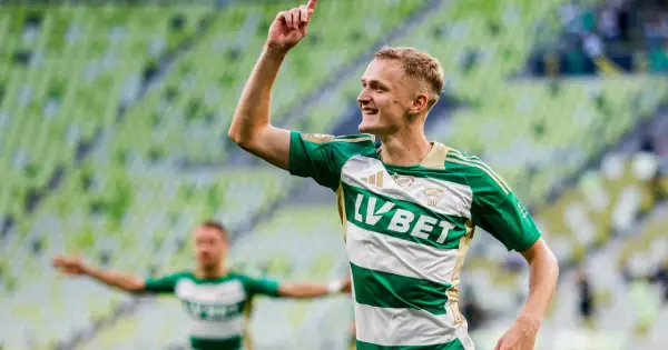 Radosny środkowy napastnik w zielono-białej koszulce świętuje bramkę na tle zielonych trybun stadionu.