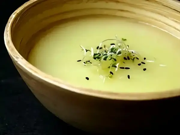 Un bol en bois contient un velouté d'asperges blanches, garni de pousses vertes et de graines noires.
