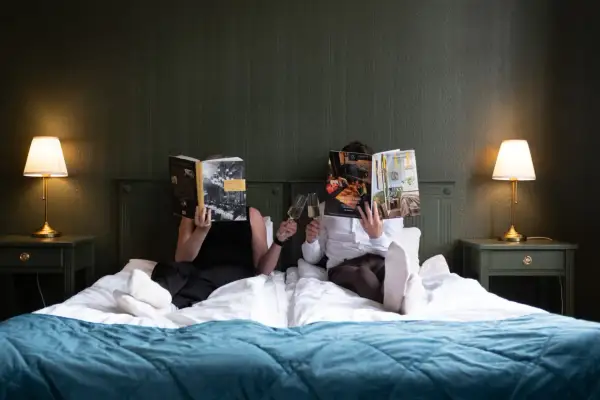 Två personer läser tidningar och skålar med champagne i en säng på Hotell Hertig Karl Filipstad.